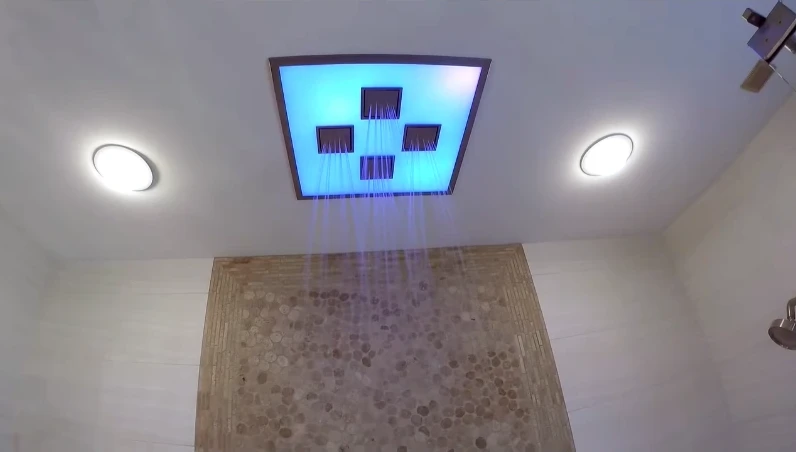 Ceiling-mounted rain shower panel with soft blue light over a pebble-tile floor