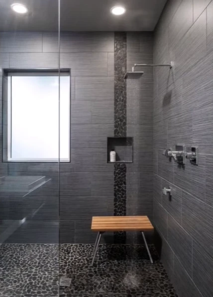 Dark gray tile shower with a vertical mosaic stripe, teak bench and pebble floor