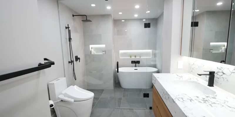 Gray tile bathroom with a glass walk-in shower, freestanding tub, lit niches and a marble vanity