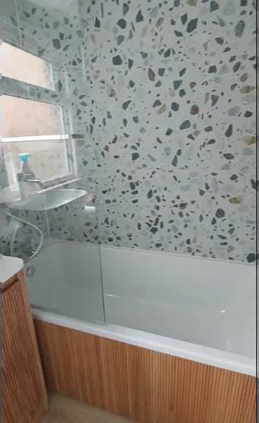Bathtub with a glass bath screen, terrazzo tile walls and a wood slat tub panel under a frosted window