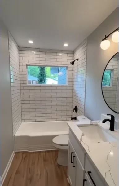 Remodeled bathroom with a white subway tile tub surround, wood-look floor, white vanity and round black mirror