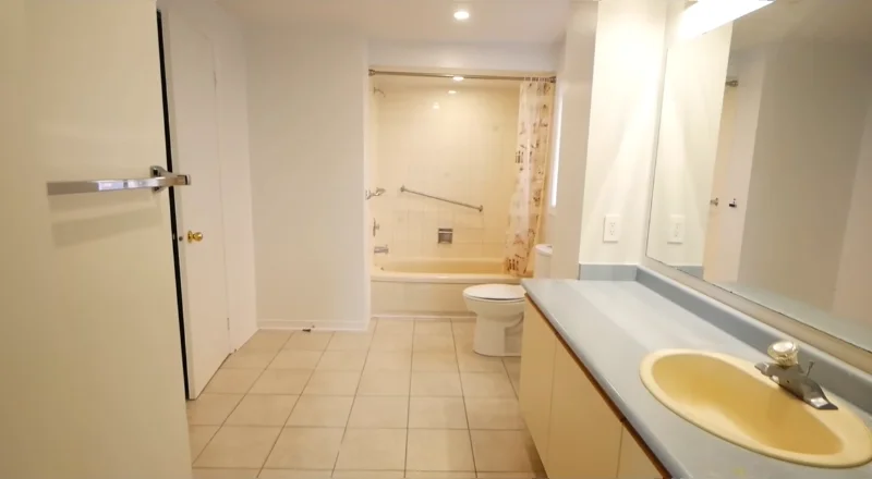 Dated bathroom with a yellow sink, laminate counter, beige floor tile and a tub at the far end before remodeling