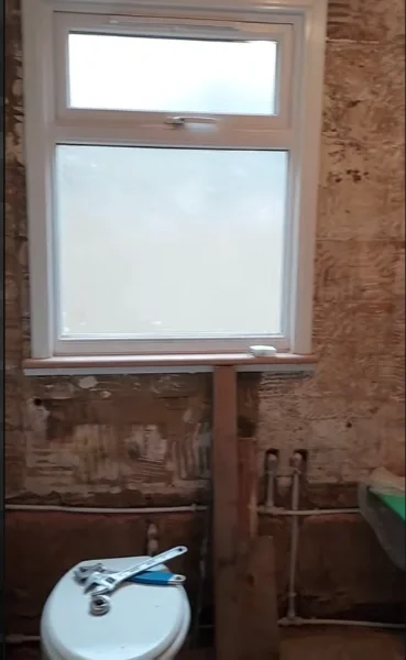 Bathroom wall stripped to exposed brick with open supply pipes below a frosted window