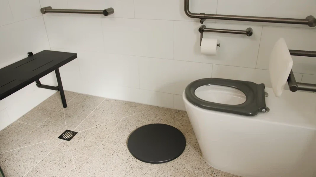 Round swivel transfer plate on a pitched speckled floor beside a floor drain, shower bench and raised toilet