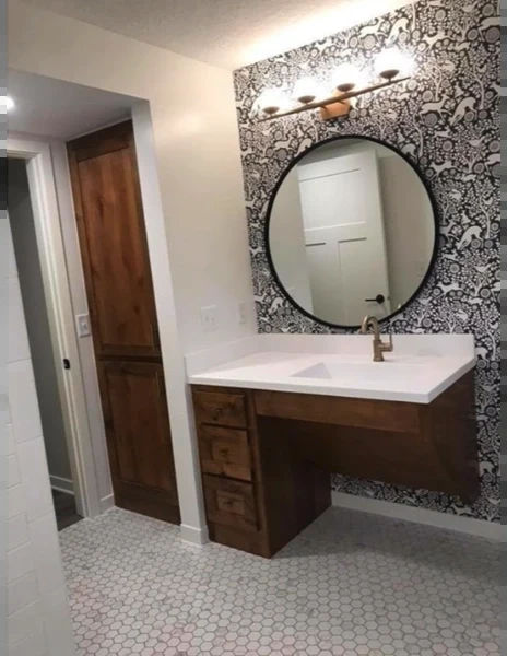 Roll-under wood vanity with a white countertop, a round black mirror and a patterned accent wall