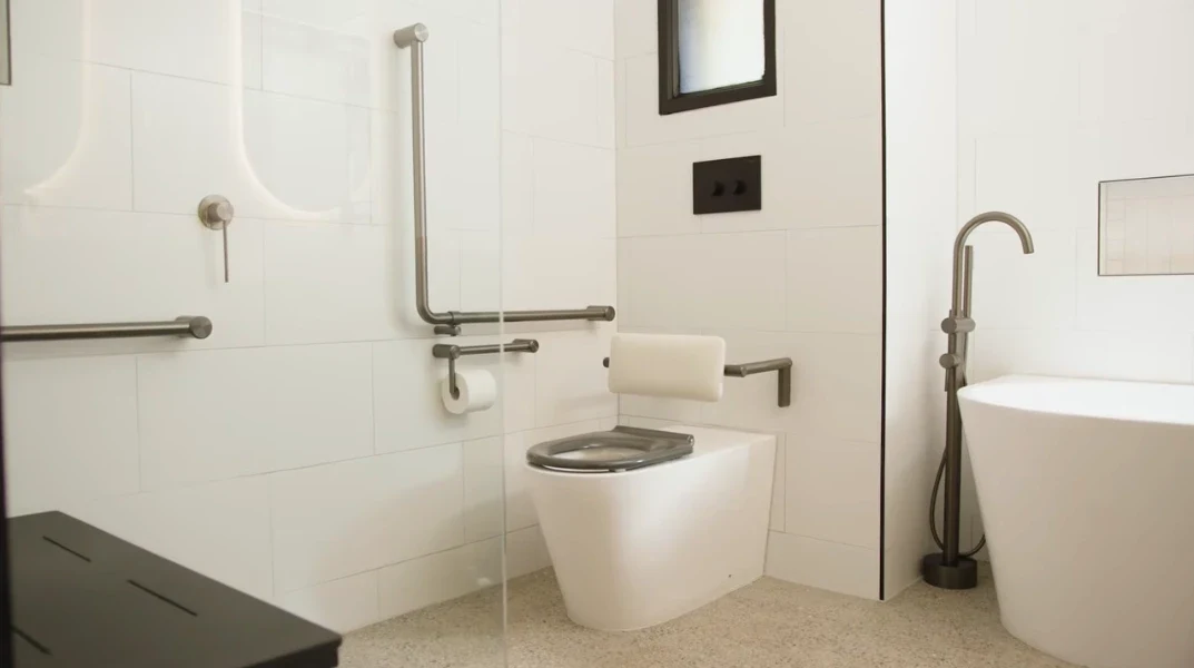 Walk-in shower with L-shaped grab rails beside a wall-hung toilet with a padded backrest and drop-down support arm