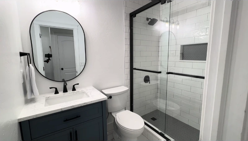 Oval black-framed mirror above a navy vanity beside a sliding glass shower door